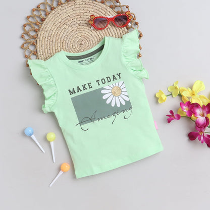 Mint green t-shirt with floral design and text, placed on a textured surface with flowers and lollipops.